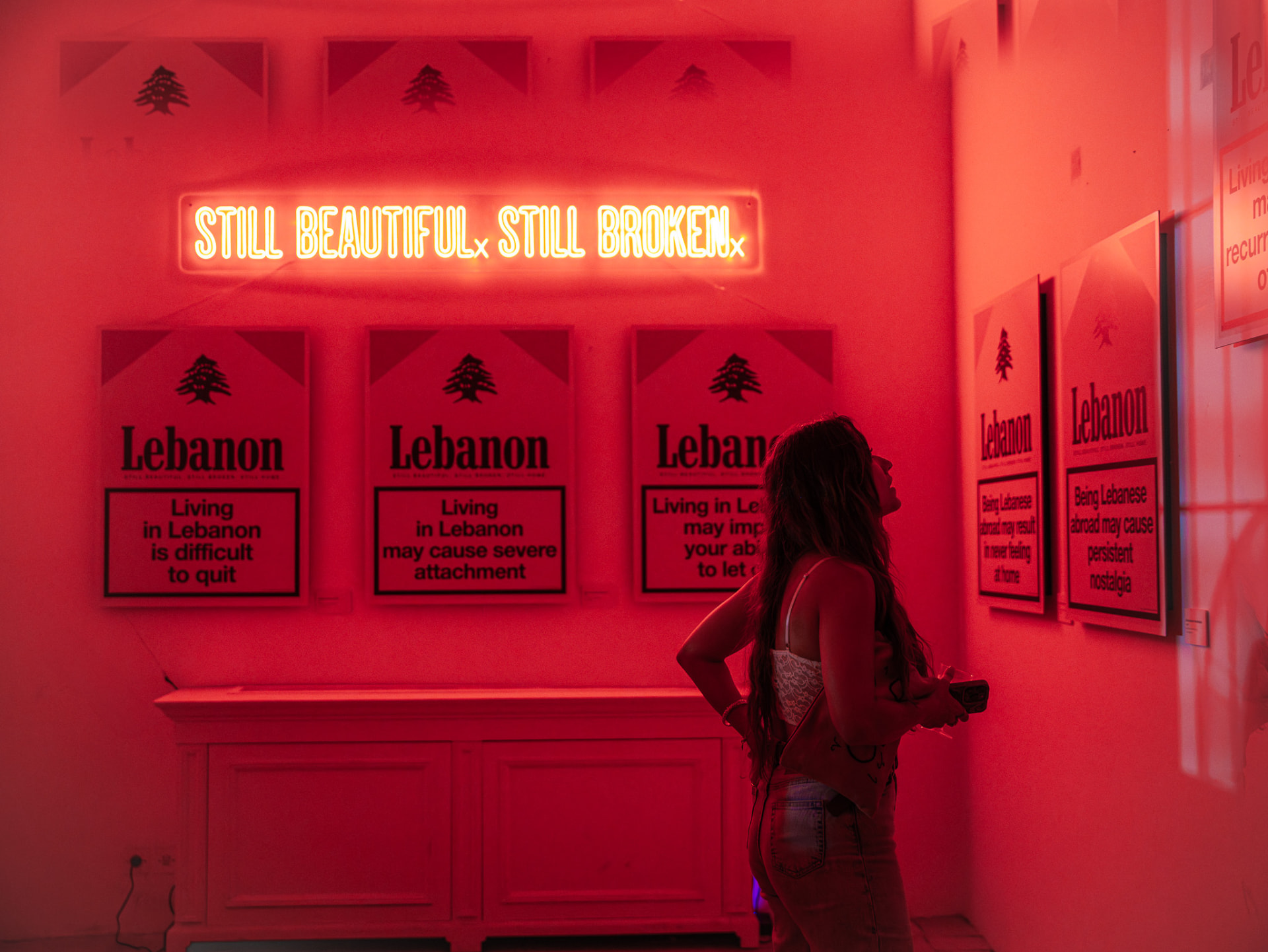 Visitor bathed in red light beneath the neon, looking up at the prints